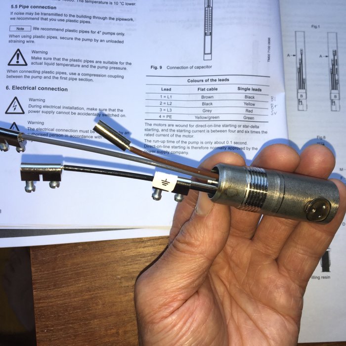 Hand holding a circular electrical connector with three screws, in front of an open manual showing wiring diagrams.