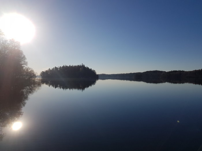 Solen reflekteras i en lugn sjö med skogklädda öar och klarblå himmel.