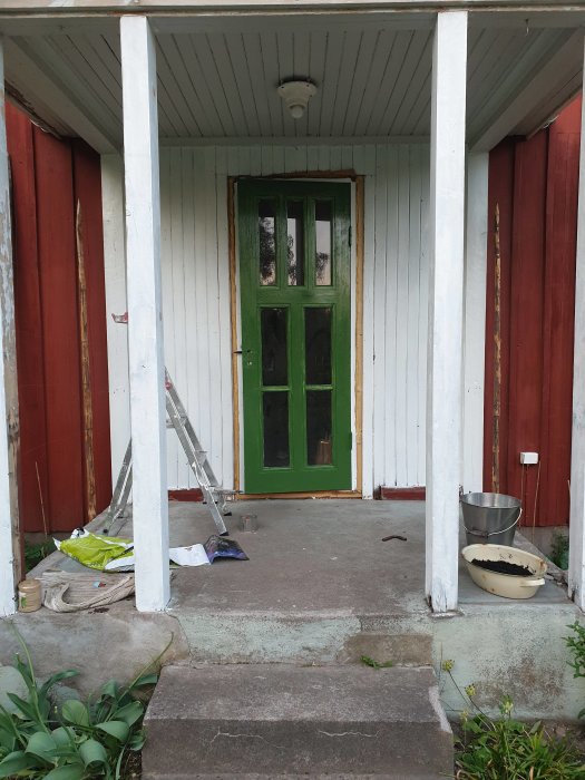 Framsidan av ett hus under renovering, grönmålad dörr, vit fasad, stege och målarmaterial framför.