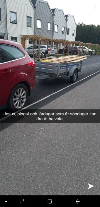 Röd bil med släpvagn full med träplank i ett bostadsområde.