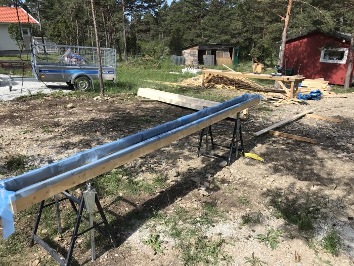 Behandlad träpanel med järnvitriol på bockar utomhus framför en röd stuga och en släpvagn på en byggplats.