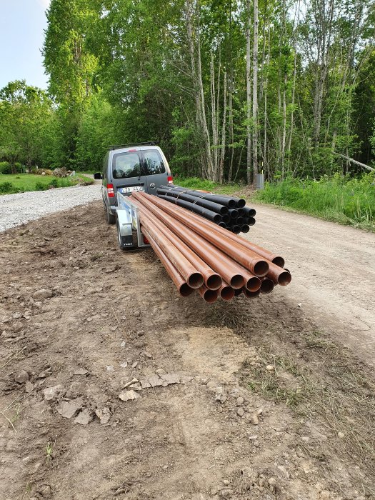 Pickup lastad med långa plaströr och metallprofil står vid en grusväg omgiven av gröna träd.