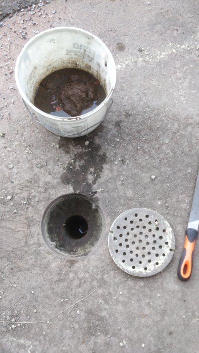 Öppen utomhusbrunn med igensatt hål, hink med grus bredvid och avtaget brunnslock.