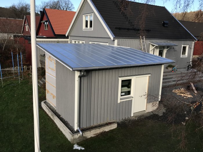 Halvfärdig grå snickarbod framför bostadshus med vita knutar och foder, soligt väder.