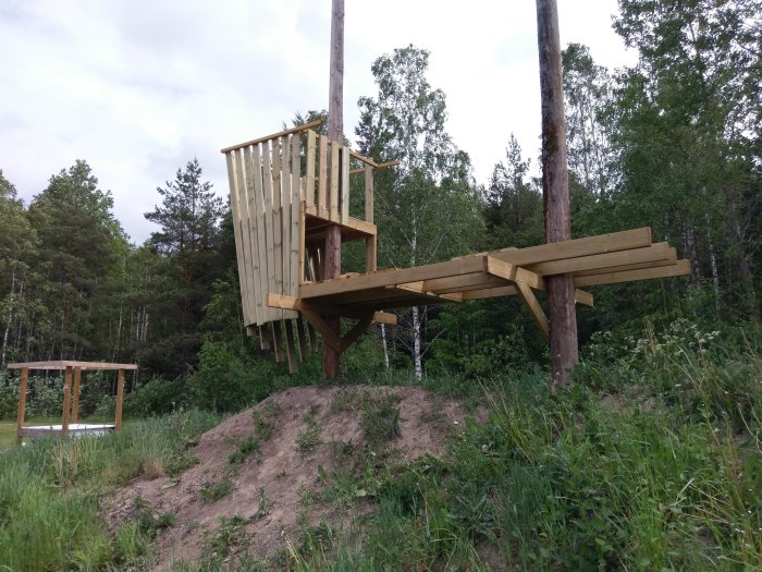 Trädäck på stolpar ovanför en kulle med svaj, omgivet av gröna träd och buskar.