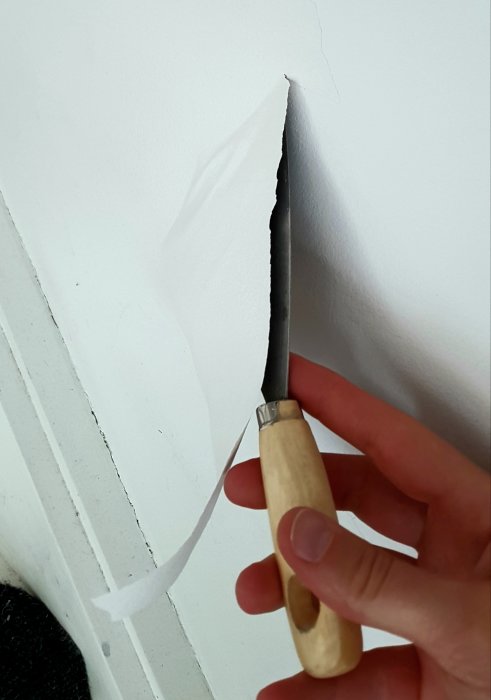 En hand som skrapar bort vit färgfilm från en vägg med en puttykniv.