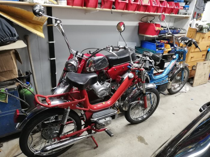 Två röda klassiska italienska motorcyklar parkerade i ett garage med en blå moped i bakgrunden.