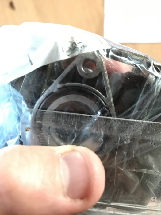 Hand håller en genomskinlig linjal framför en förgasare inuti en plastpåse för att mäta komponentens storlek.