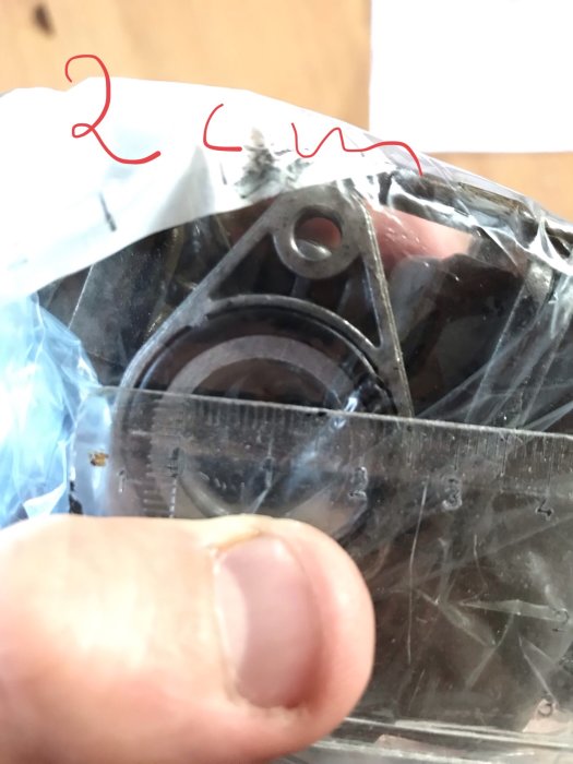 Någons hand håller en transparent linjal mot en maskindel i en plastpåse, med röd text "2 cm" på påsen.
