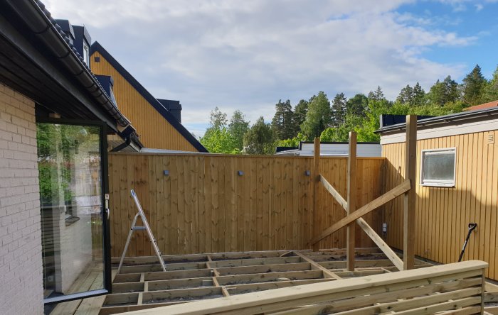 Nybyggd veranda under konstruktion med trästomme och stolpar, nära ett hus och staket.