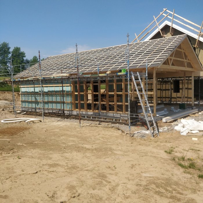 Ett garage under konstruktion med takstolar uppe och byggmaterial på marken under en klarblå himmel.