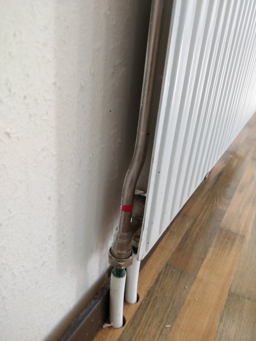 Radiator ansluten till väggen utan synliga avstängningsventiler vid anslutningsrören på trägolvet.