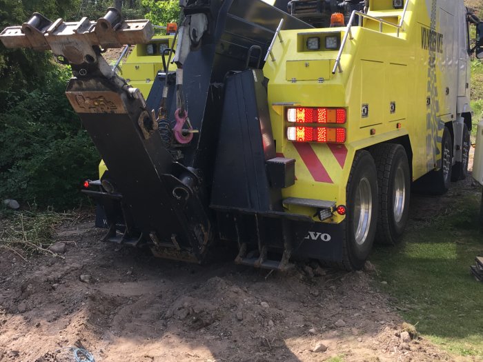 Gul lastbil med lyftanordning påbörjar lyft av stor sten, omgivet av grönska.