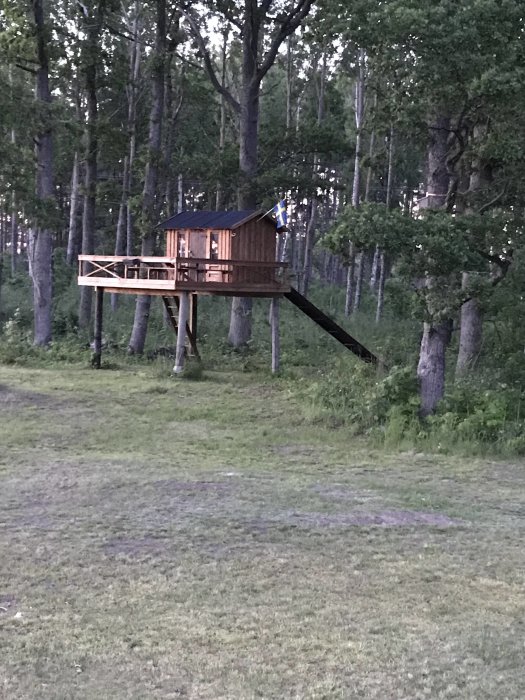 Lekstuga byggd på höga stolpar med stödben i en skogsglänta, flagga på taket, kvällsljus.