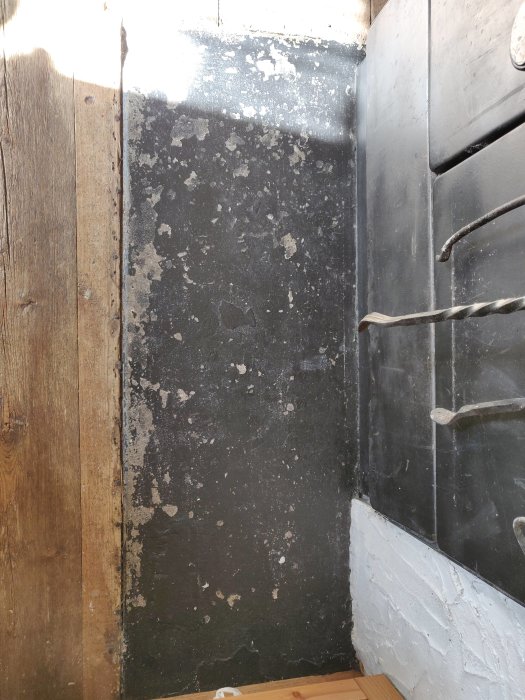 Slitet trägolv med färgfläckar, gropar och springor bredvid en svartmålad vägg och vitmålad tröskel.