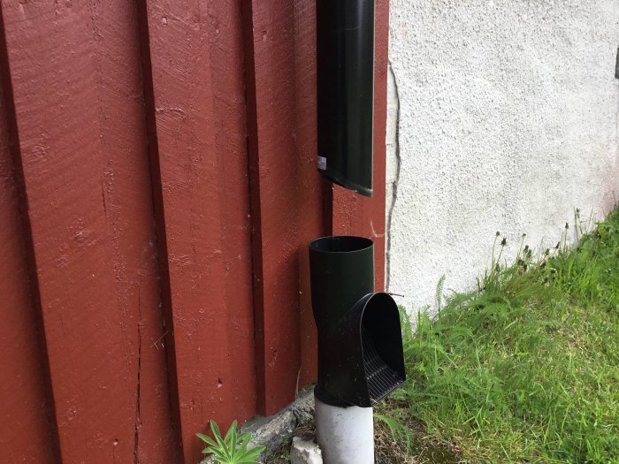 En delvis blekt dräneringsrör vid en röd husvägg bredvid en vit putsad yta.