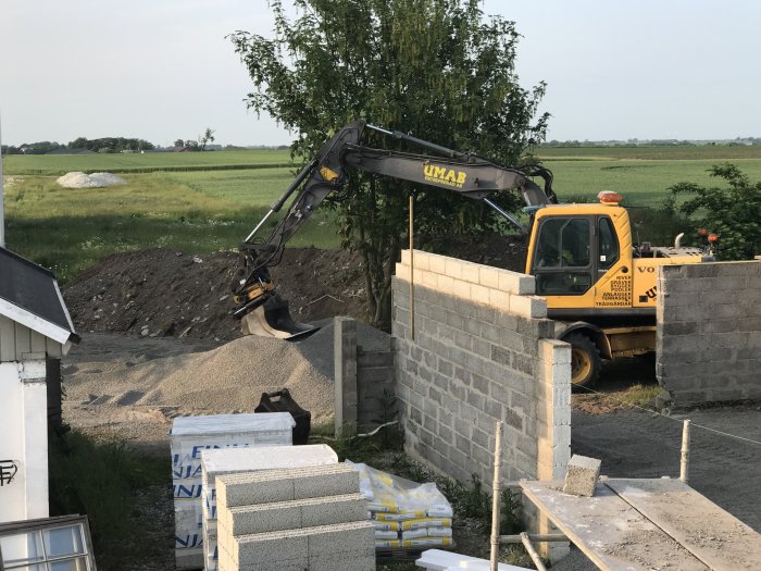 Grävmaskin och byggmaterial vid halvfärdigt garage med lecaväggar, i landsbygdsmiljö.