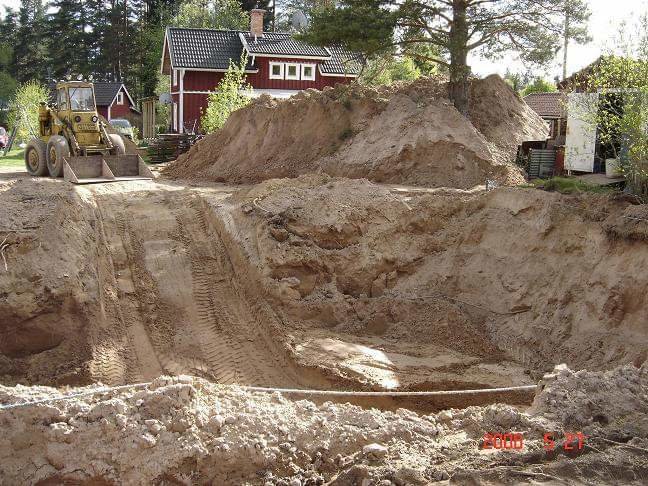 Hög med fyllnadsmassor vid villa med lastmaskin och grävd grop i trädgården.