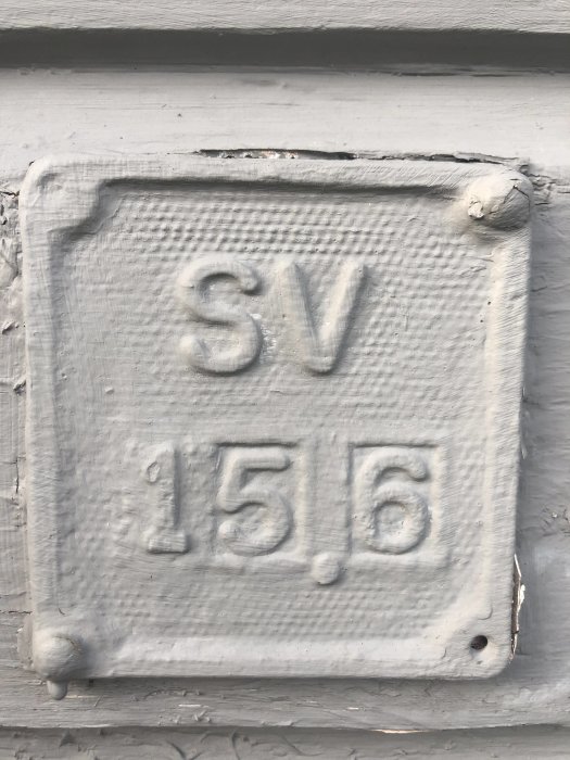 Gråmålad skylt på vägg med texten "SV 15,6" i relief.