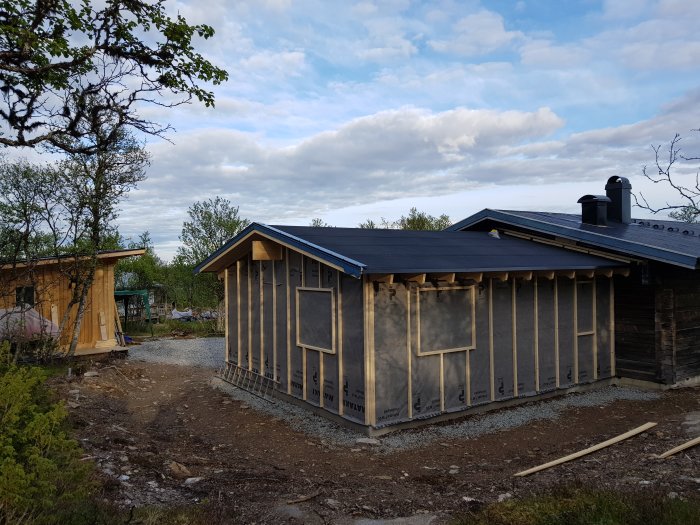 Nybyggd stuga med takplåt och pågående arbete med vindduk och spikläkt, omgiven av natur.