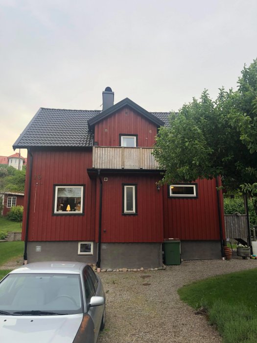 Rödfärgat hus med svart tak och synlig skorsten, parkerad bil framför, omgiven av grönska och grusuppfart.