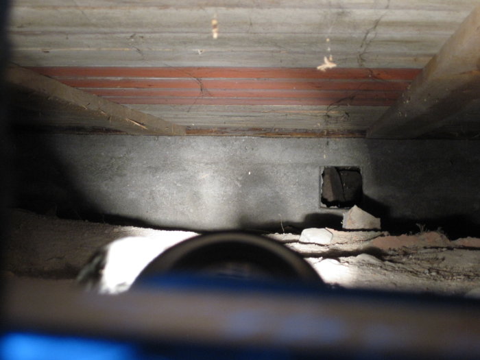 Utsikt under golv i äldre timmerhus med synlig blindbotten och grundmur, fotograferat med blixt i begränsat utrymme.