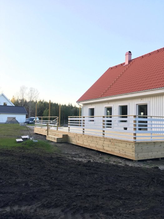 Ett luftigt vita räcket runt en träterrass på ett hus med rött tak, med utsikt över en trädgård och skog.