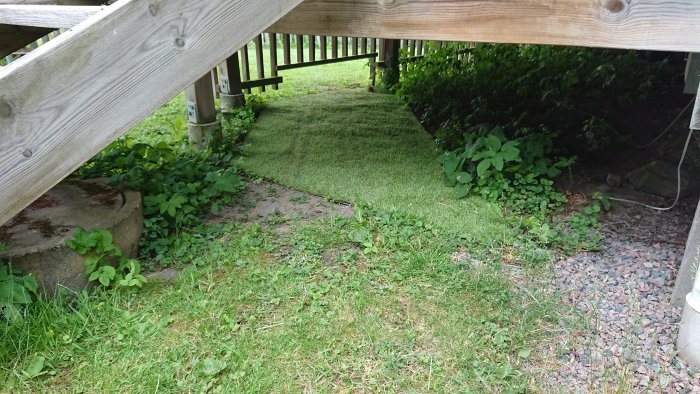 Konstgräsbelagd passageway under en träaltan med grus och vegetation runtomkring.