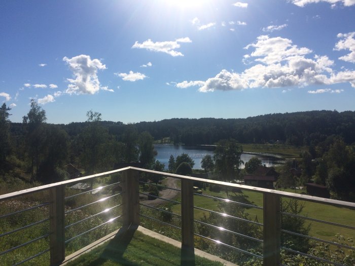 Rostfritt räcke med horisontella rör framför en utsikt över en sjö, gröna träd och klar himmel.