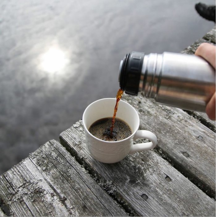 Hälla bryggkaffe från termos i vit kopp på träbrygga med vatten i bakgrunden.