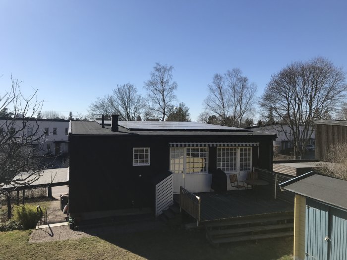 Ett hus med ett svart platt tak utrustat med stora solpaneler, ny takpapp och borttagna genomföringar, omgivet av träd och en klarblå himmel.