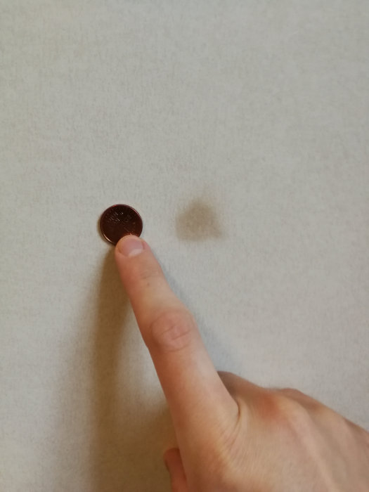 En hand som pekar på en liten mörk fläck på en ljus väggyta med en enkrona för storleksskala.