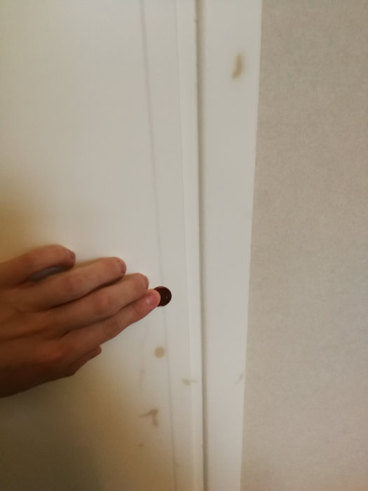 Hand håller en enkrona mot en vit dörrkarm med bruna fläckar för storleksjämförelse.
