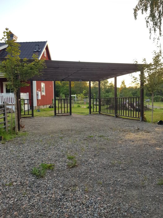 Träpergola med tak som fungerar som välkomstentré framför ett rött hus, omgivet av grus och grönska.