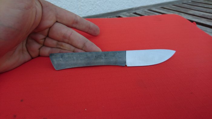 Hand håller en delvis slipad fickjaktkniv med 2.6 mm tjockt RWL 34 blad mot röd bakgrund.