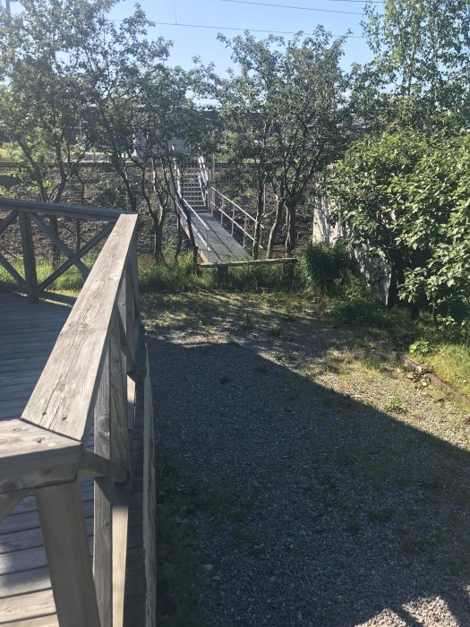 Nybyggd gångbro som leder till en tomtgräns omgiven av grönska under en solig dag.