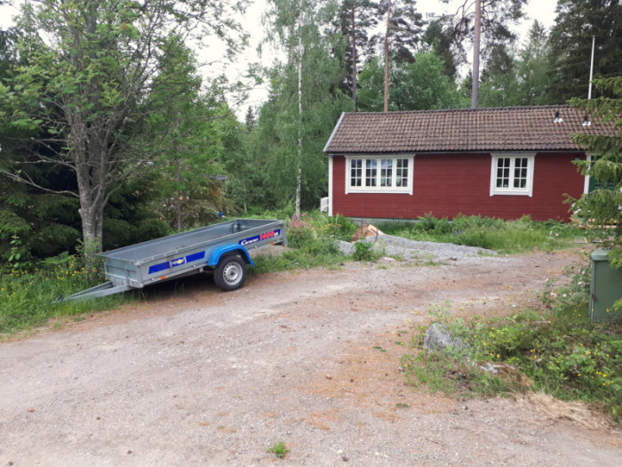 En bild på en uppfart med en släpvagn framför ett rött hus med vita knutar, trädbädd och skog i bakgrunden.