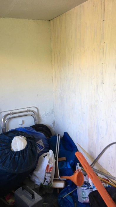 En oorganiserad garagehörn med en vägg där färgen har flagnat och lossnat, omgiven av olika föremål som säckar och rengöringsredskap.