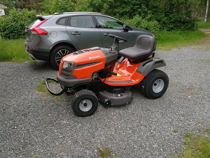 Ny trädgårdstraktor parkerad på grus framför en grå bil, omgiven av grönska.