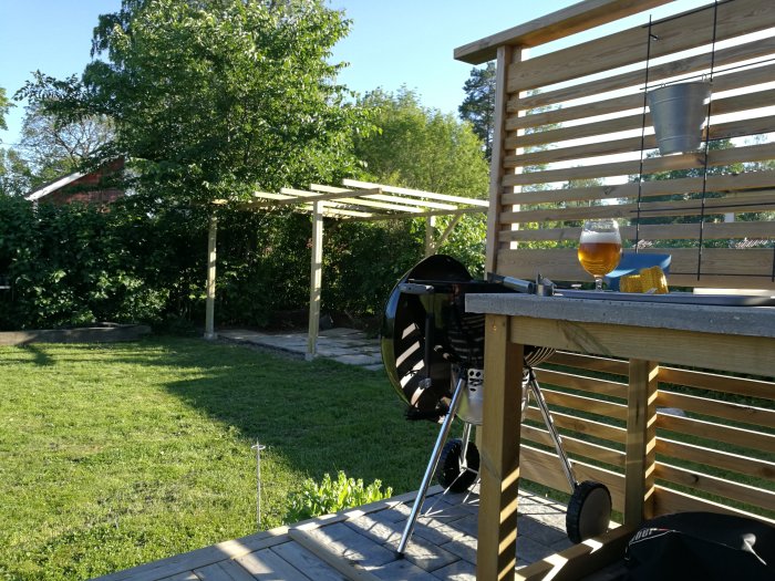 Nybyggd pergola av trä i en trädgård med grill och glas på bordet, gammal marksten synlig.