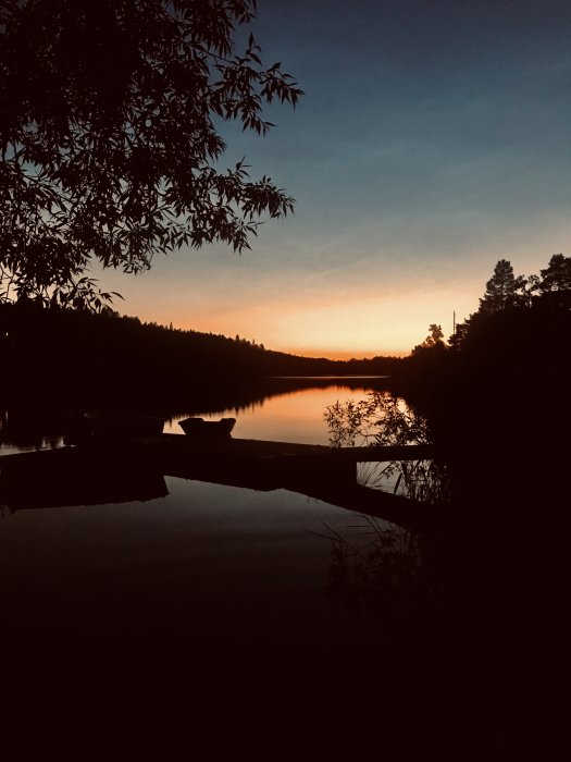 Solnedgång över en stilla sjö med trädens silhuetter och en båt vid en brygga.