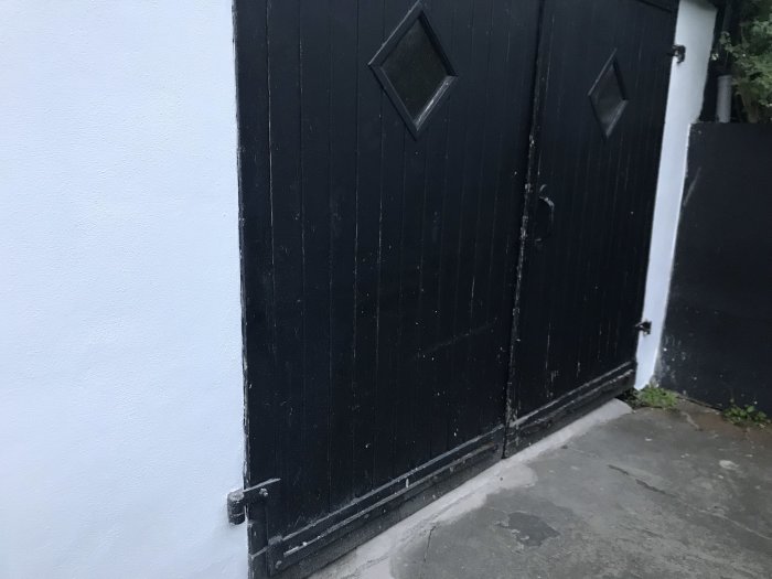 Gammal svart garagedörr med järnbeslag, övre delen fin, nedre delen skadad av väder och slitage.