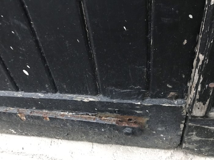 Nedre delen av en gammal svart garagedörr med rost och skador från regn och slitage.