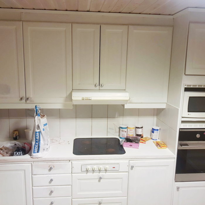 Gammal vit köksfläkt ovanför spishäll i ett ljust kök med vit inredning och spretiga ingredienser på bänken.