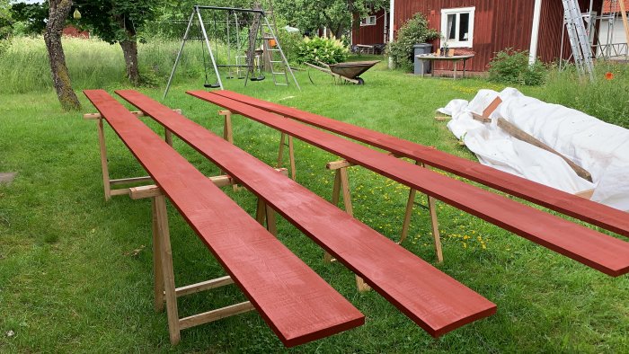 Rödmålade lockbrädor på bockar i en trädgård med hus och stege i bakgrunden.