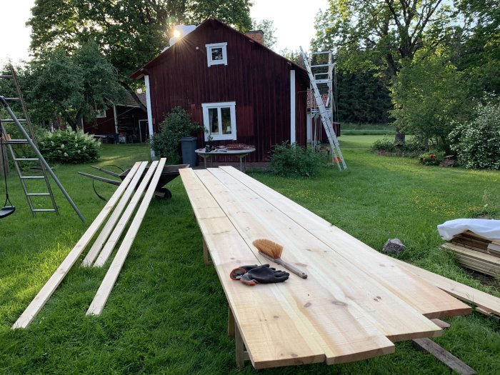 Träbrädor på gräset med hus och stege i bakgrunden, byggarbete pågår.