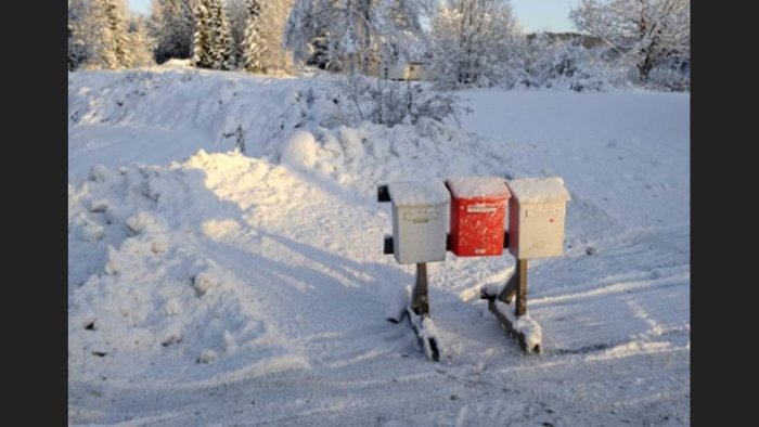 Två brevlådor på ställning vid en snötäckt vägkant med vinterlandskap i bakgrunden.