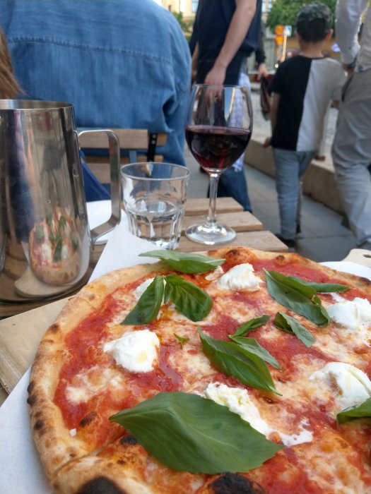 Pizza med tomatsås, mozzarella och färska basilikablad på ett bord utomhus med ett glas vin och vatten.