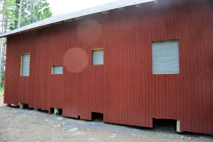 Rödmålad träfasad på fritidshus med takfot och ofärdiga fönsteröppningar där isolering syns.