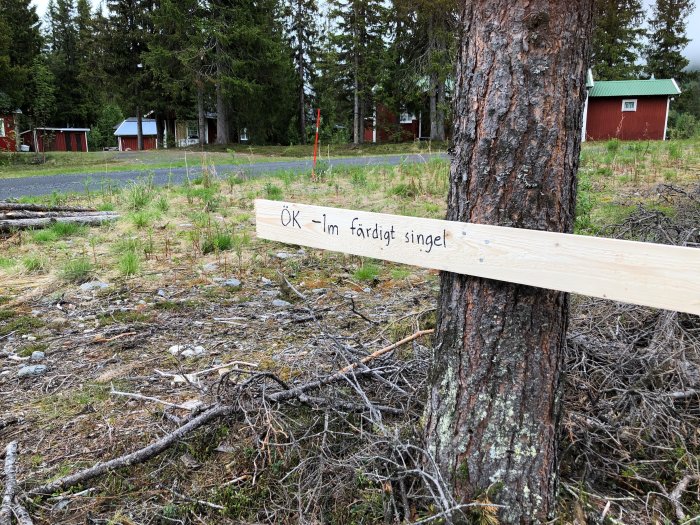 En skogstomt med markerad plats för stuggrund, en trälist med texten "OK -1m färdigt singel" mot en trädstam, i bakgrunden syns röda stugor.